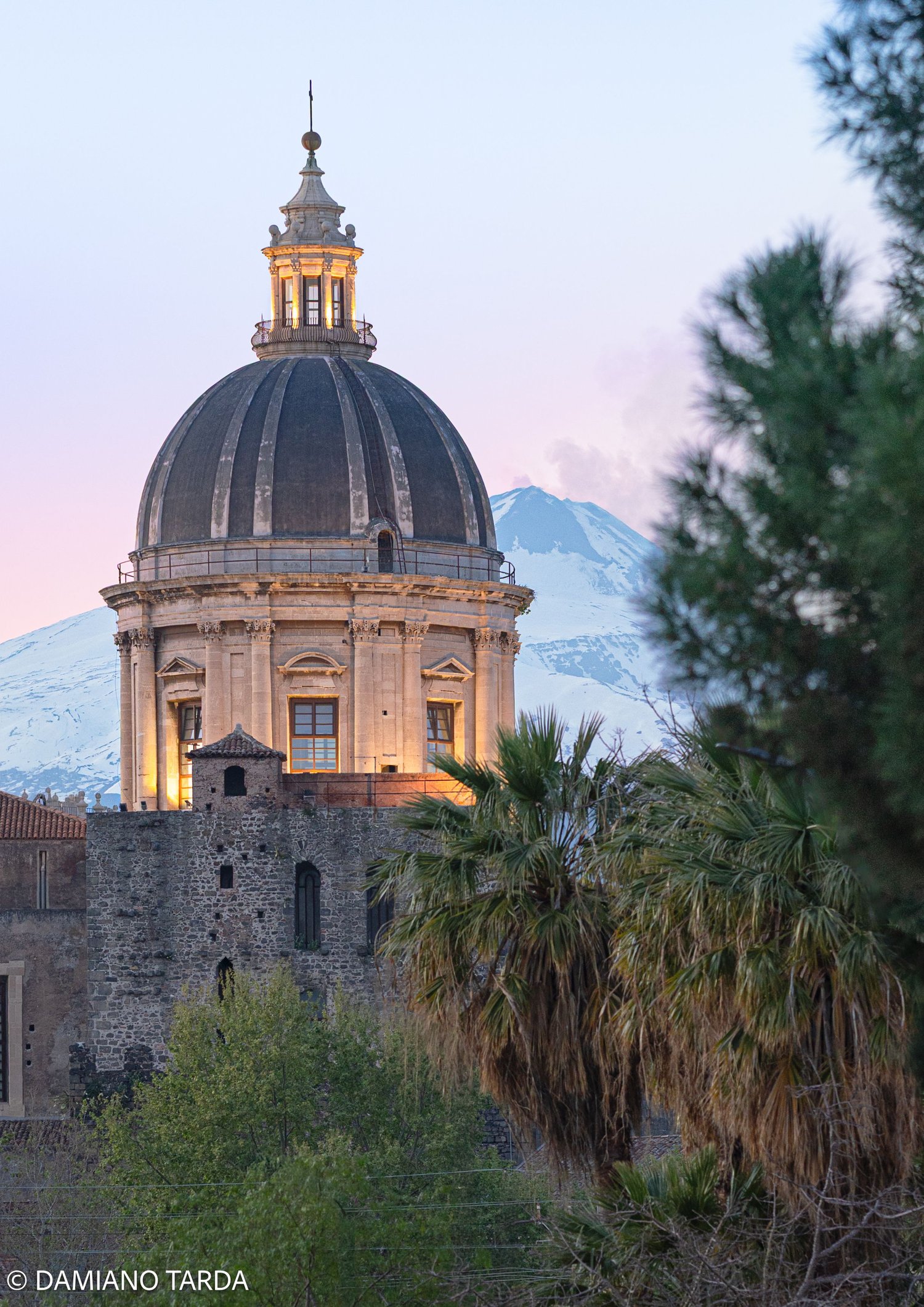Dome of a Baroque church in Catania at sunset, with snow-capped Etna in the background.